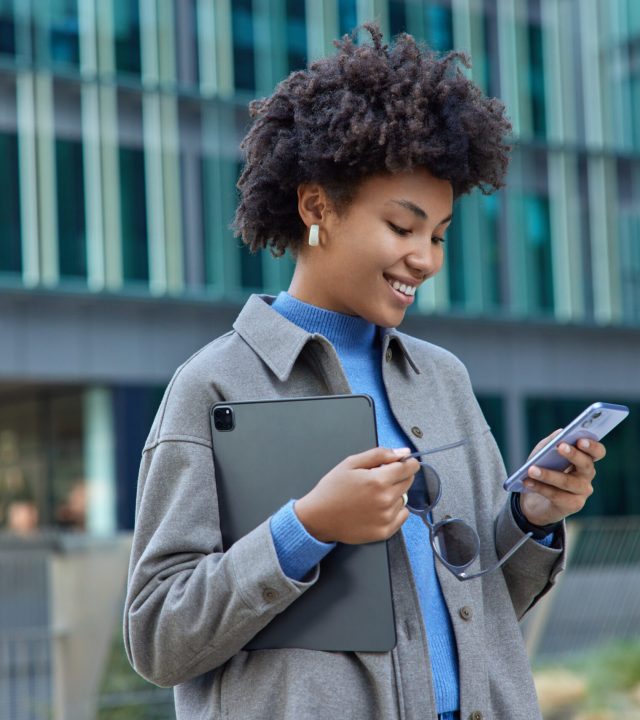 Waist up shot of happy curly haired woman uses modern technologies in city holds mobile phone and digital portable tablet smiles gladfully as reads pleasant text message wears stylish clothes