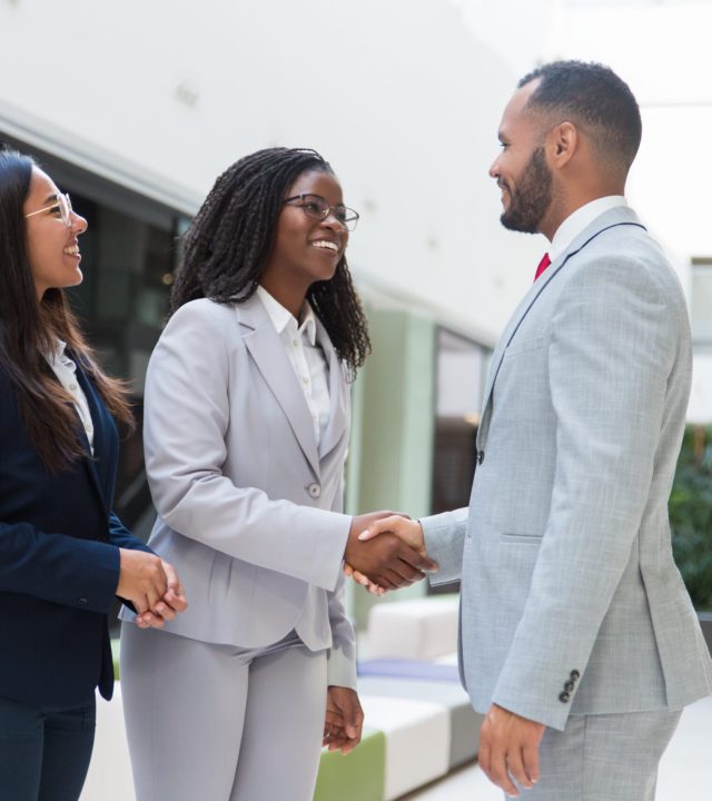 Happy joyful diverse business partners greeting each other in hallway. Business man and women standing, shaking hands with each other, talking and smiling. Successful partnership concept